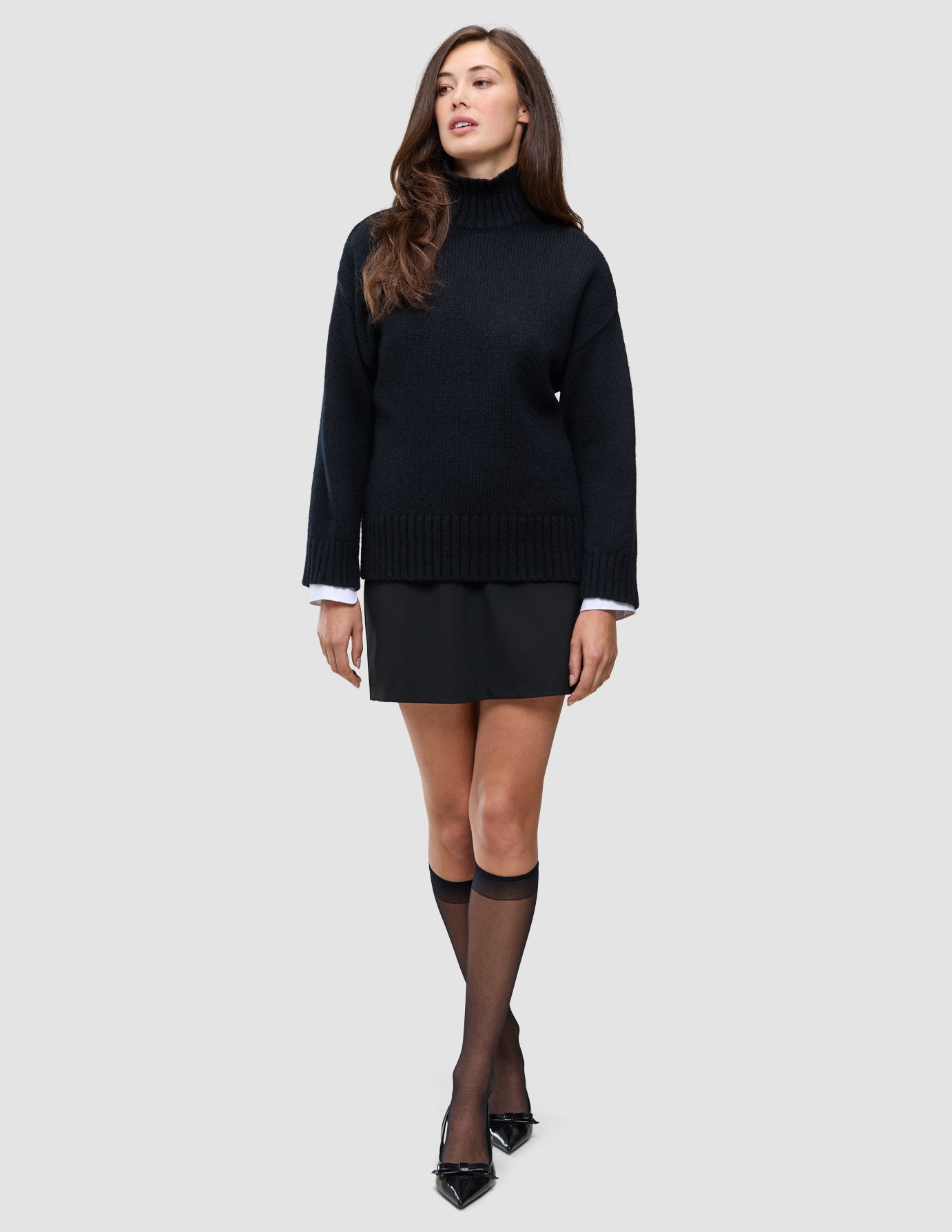 En kvinne har på seg en svart, tykk og myk ullstrikket turtleneck for kvinner over en hvit skjorte med krage, et svart skjørt og svarte høyhælte sko.